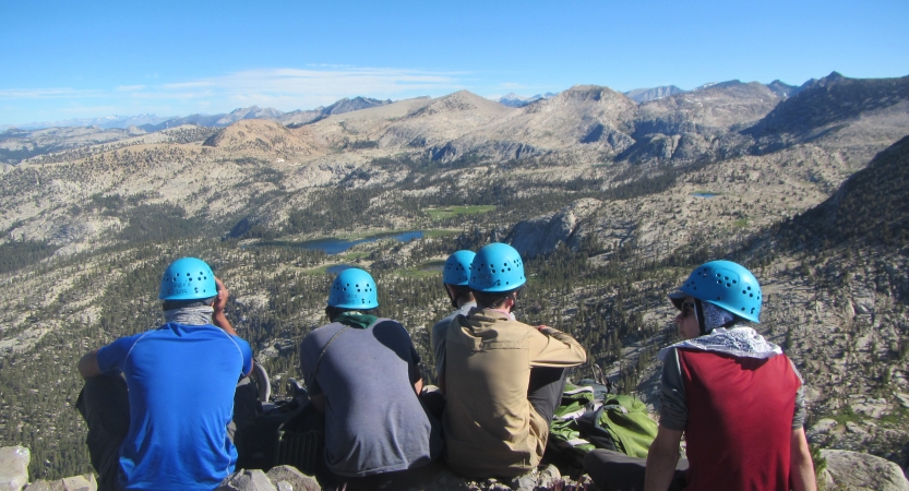 Five people wearing helmets stand atop a rocky summit overlooking mountains. 